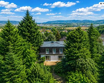 Luftaufnahme eines Hauses, umgeben von üppigem Grün und hohen Bäumen, mit Fernblick auf die Landschaft.