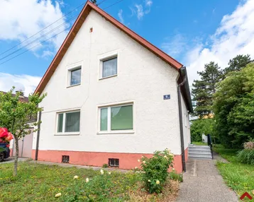 Vorderansicht eines Einfamilienhauses mit weißer Fassade und rotem Dach in einer grünen Umgebung.