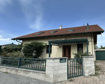 Hausfassade mit Vorgarten, Zaun und Einfahrt unter blauem Himmel.