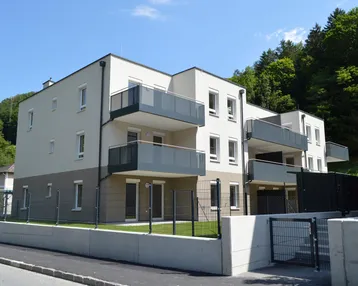 Modernes Mehrfamilienhaus mit Balkonen und kleinen Gärten, umgeben von grüner Natur.