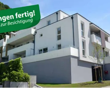Modernes Mehrfamilienhaus mit weißer Fassade, Balkonen und grüner Umgebung am Hang.