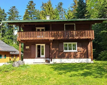 Traditionelles Holzhaus mit Balkon und grüner Umgebung, umgeben von hohen Bäumen.