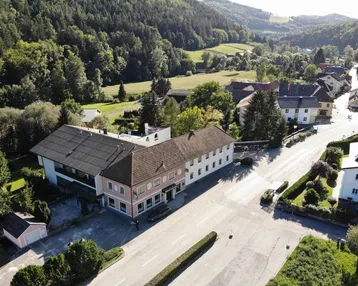Luftaufnahme eines Dorfes mit einem großen Gebäude und umliegender grüner Hügellandschaft.