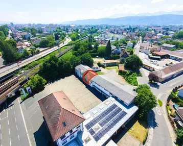 Luftaufnahme eines Gewerbeobjekts mit mehreren Gebäuden, großem Parkplatz und Solaranlage auf dem Dach.