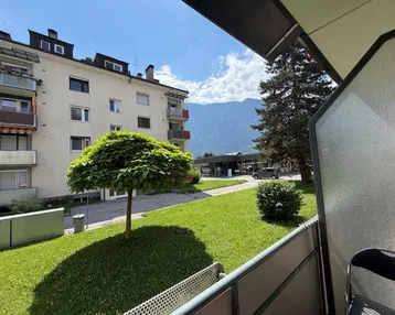 Blick vom Balkon auf die grüne Umgebung, ein Mehrfamilienhaus und die Berge im Hintergrund.