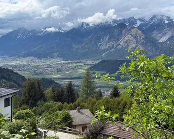 Panoramablick auf die umliegende Berglandschaft und das Tal mit vereinzelten Häusern unter bewölktem Himmel.