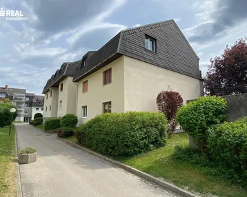 Mehrfamilienhaus mit mansardähnlichem Dach und gepflegter Grünanlage unter bewölktem Himmel.
