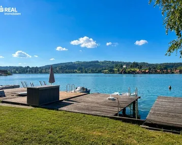 Idyllischer Blick auf den See mit privatem Holzsteg und Liegeflächen unter blauem Himmel.
