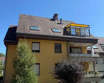 Gelbe Fassade eines Mehrfamilienhauses mit Balkonen und Dachfenstern unter blauem Himmel.