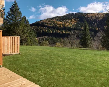 Rasenfläche mit Holzzaun und Blick auf bewaldete Berge unter blauem Himmel.