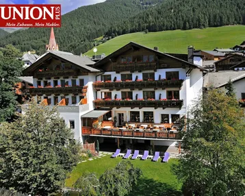Großzügiges Hotelgebäude mit traditioneller Architektur, Balkonen, Gartenbereich und Bergblick in einer malerischen Umgebung.