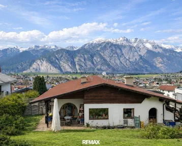 Haus mit rotem Dach und Bergblick in ländlicher Umgebung unter blauem Himmel.