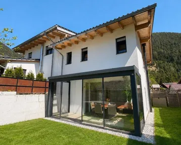 Modernes Haus mit verglastem Wintergarten und gepflegtem Rasen, umgeben von grüner Natur.