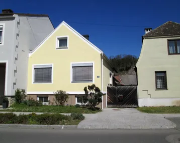 Gelbes Einfamilienhaus mit Satteldach und Rollläden an den Fenstern, umgeben von einem kleinen Vorgarten.
