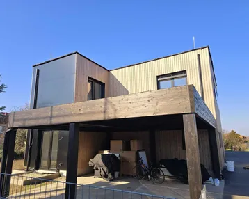 Modernes Haus in Holzbauweise mit Carport und Baustellencharakter unter blauem Himmel.