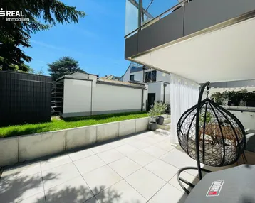 Große, sonnige Terrasse mit Fliesenboden und Hängesessel, umgeben von Grünflächen.