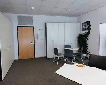 Geräumiges Büro mit Teppichboden, weißen Schränken und einem kleinen Besprechungsbereich für Mitarbeiter.