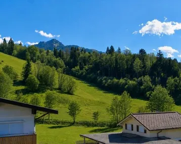 Panoramablick auf grüne Hügel und bewaldete Berge mit traditionellen Häusern unter blauem Himmel.