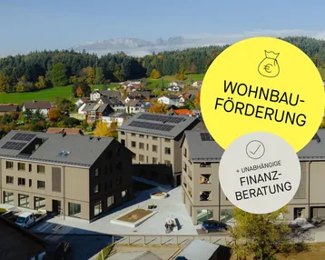 Luftaufnahme von modernen Wohngebäuden mit Solaranlagen und Bergkulisse, umgeben von grüner Landschaft.