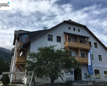 Mehrfamilienhaus mit traditioneller Architektur, Holzbalkonen und einem kleinen Spielplatz im Vordergrund.