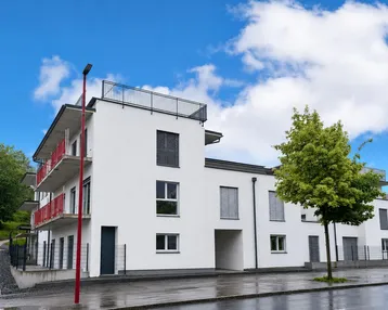 Modernes Mehrfamilienhaus mit weißer Fassade, roten Balkonen und Flachdach unter blauem Himmel.