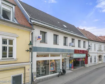 Fassade eines Geschäftsgebäudes mit Schaufenstern und angrenzenden historischen Gebäuden an einer Straße.