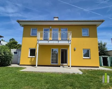 Gelbes Einfamilienhaus mit Balkon, kleiner Terrasse und grünem Garten unter blauem Himmel.