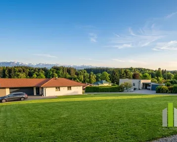 Weitläufige Landschaft mit einem modernen Haus, umgeben von grünen Wiesen und einem beeindruckenden Bergblick.