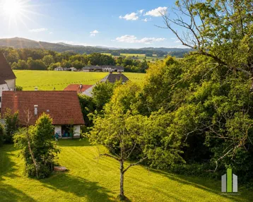 Panoramablick auf ein Haus mit rotem Ziegeldach, umgeben von grünen Wiesen und Bergen unter strahlendem Himmel.