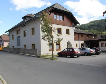 Gebäude mit traditioneller Architektur, Holzverkleidung im Dachbereich und Parkplätzen davor, umgeben von grüner Landschaft.