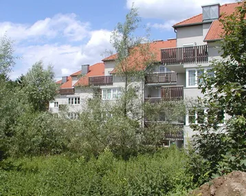 Mehrfamilienhaus mit Balkonen und roten Ziegeldächern, umgeben von üppigem Grün und Bäumen.