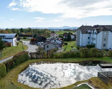 Panoramablick auf die ländliche Umgebung mit einem Teich, Wohnhäusern und fernen Bergen.