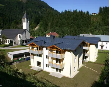 Neubauwohnungen mit Balkonen und Bergblick, umgeben von grünen Wäldern und einer Kirche.