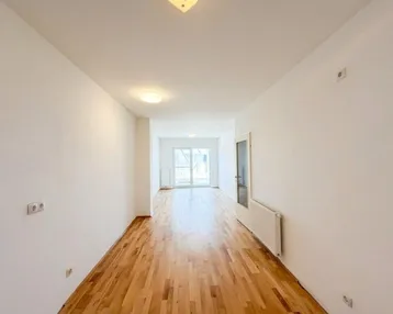Helles, leeres Zimmer mit Parkettboden und Zugang zu einem Balkon oder einer Terrasse.