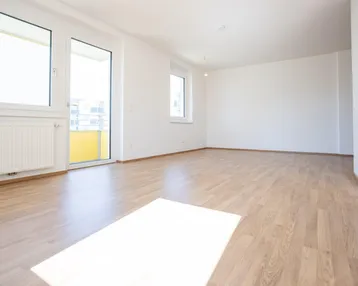 Helles, leeres Zimmer mit Laminatboden, großen Fenstern und Zugang zum Balkon.