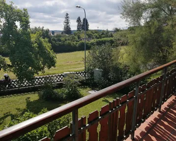 Blick vom Balkon auf eine weite grüne Wiese und bewaldete Hügel unter einem bewölkten Himmel.