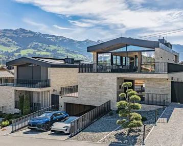 Moderne Villa mit Steinfassade, großen Fenstern und Bergblick, umgeben von grüner Landschaft.