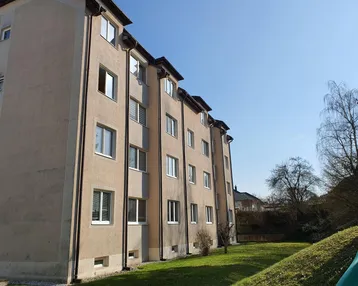 Mehrfamilienhaus mit beigefarbener Fassade und gepflegter Rasenfläche unter blauem Himmel.