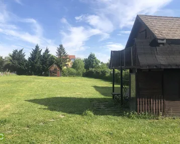 Weitläufige Rasenfläche mit einem kleinen Holzhaus und Bäumen unter blauem Himmel.
