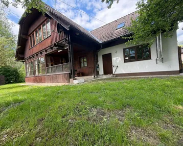 Hausfassade mit Holzverkleidung und weißem Putz, umgeben von grünem Rasen unter blauem Himmel.
