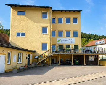 Gelbes Mehrfamilienhaus mit mehreren Etagen, Außentreppe und Parkplätzen im Vordergrund.