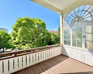 Heller Balkon mit Holzboden, weißem Geländer und Blick ins Grüne unter blauem Himmel.
