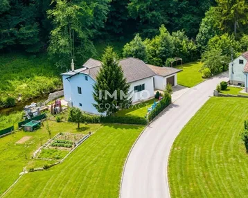 Luftaufnahme eines Hauses mit großem Garten, Zufahrt und angrenzendem Bachlauf in grüner Umgebung.