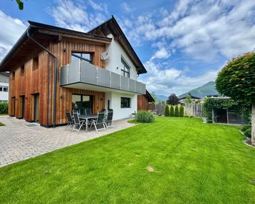 Modernes Haus mit Holzfassade, gepflegtem Garten, Terrasse und Bergblick unter blauem Himmel.