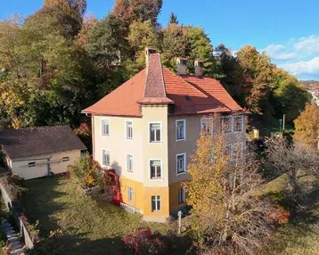Historisches Gebäude mit markantem Turm und rotem Ziegeldach, umgeben von herbstlicher Vegetation.