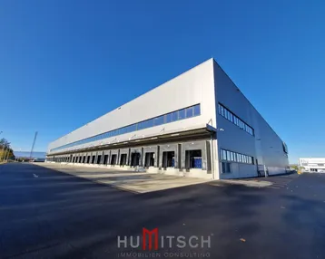 Großes modernes Industriegebäude mit zahlreichen Laderampen unter klarem blauem Himmel und weitläufigem Vorplatz.