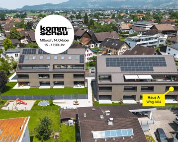 Luftaufnahme von zwei modernen Mehrfamilienhäusern mit Photovoltaikanlagen, umgeben von Grünflächen und einem Spielplatz, mit Bergblick im Hintergrund.