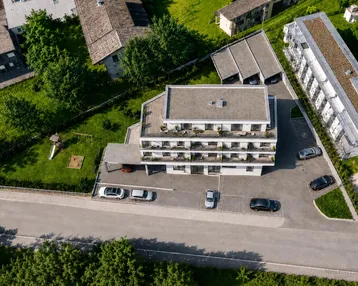 Luftaufnahme eines modernen Mehrfamilienhauses mit Flachdach, Balkonen, Parkplätzen und Grünflächen mit Spielplatz.