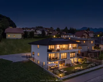 Moderne Wohnanlage bei Dämmerung mit beleuchteten Balkonen und Gärten in ländlicher Umgebung.