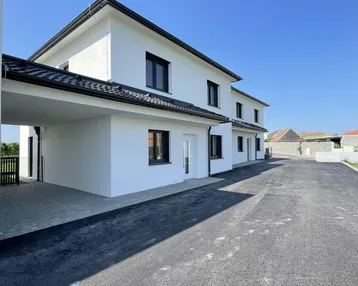Modernes weißes Gebäude mit schwarzem Dach und Fenstern, einem Carport und asphaltierter Zufahrt unter blauem Himmel.